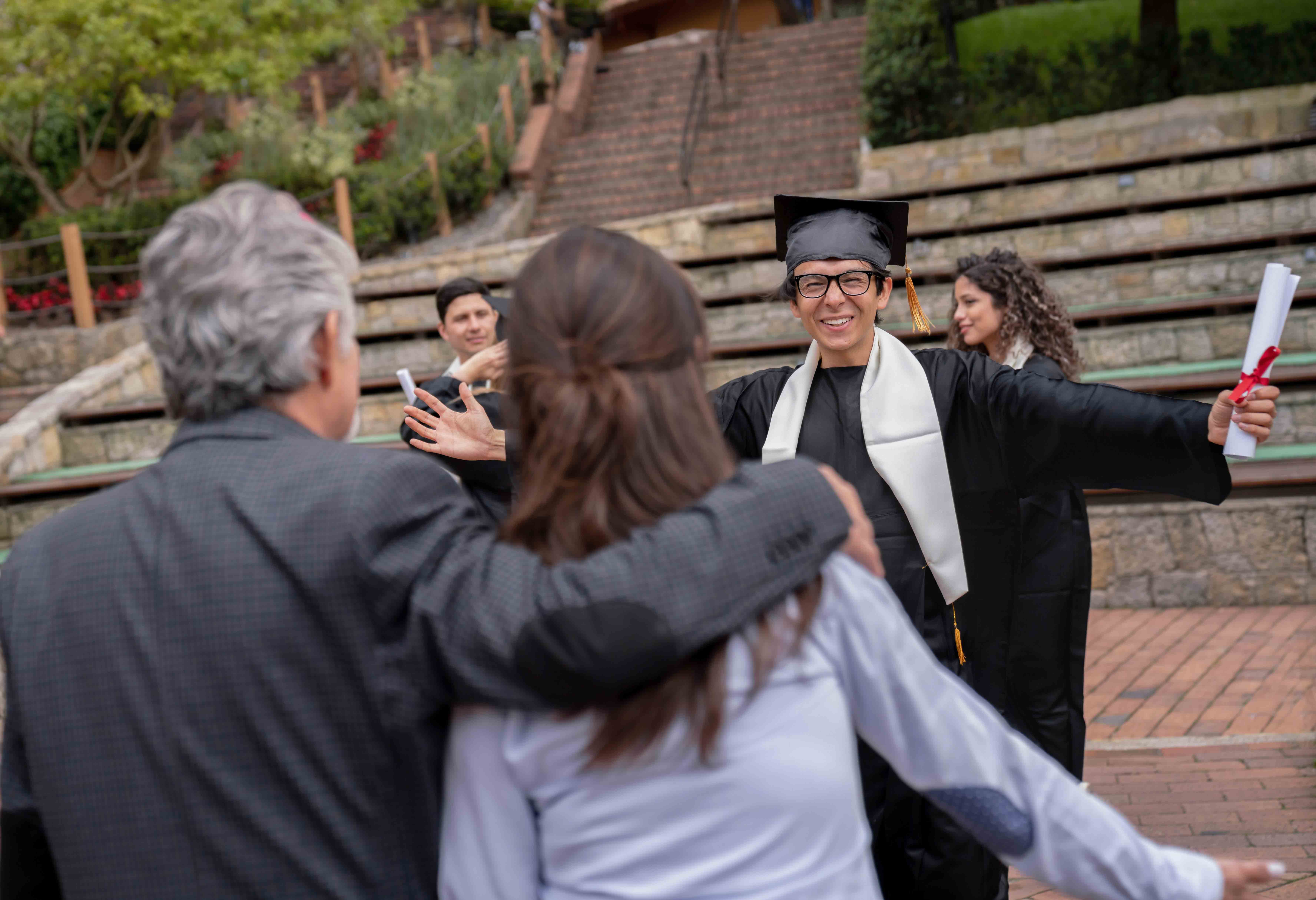 graduate wearing cap and gown with diploma, and greeting his parents